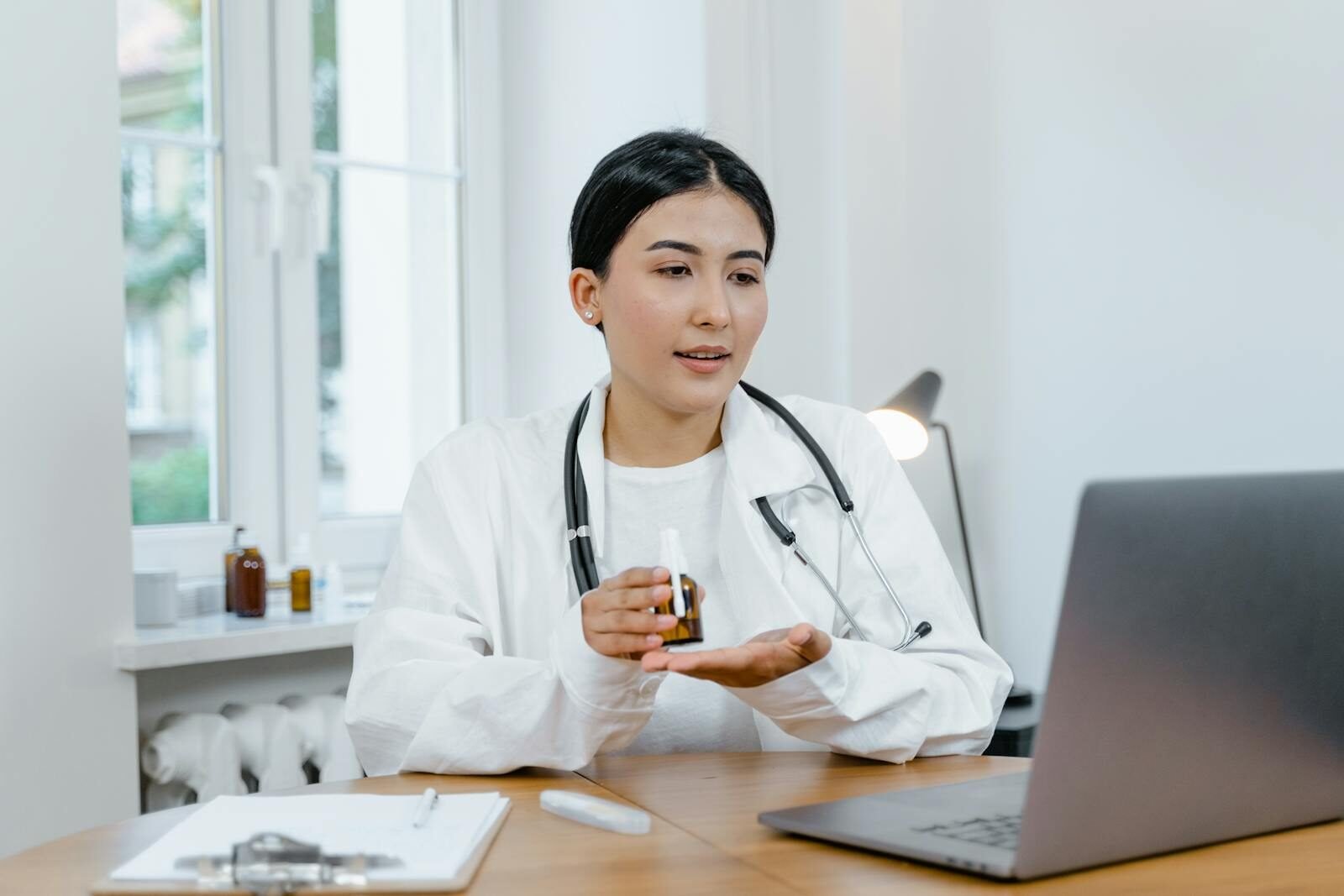 A young female doctor in a white coat on a video call, discussing medication with a patient.